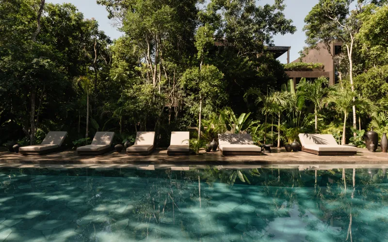 An expansive outdoor lounge area at La Reserva Tulum, featuring dark walls, built-in seating with plush cushions, natural tree trunks integrated into the design, woven pendant lights, large decorative vases, and a green patterned rug, all set agai...