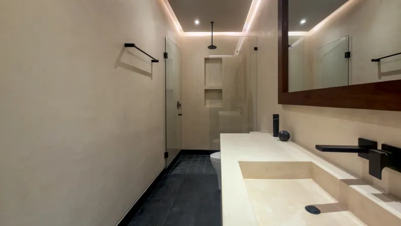 A wide shot of the open-concept dining area and kitchen in the Mestiza 15 showroom, featuring a long dark granite kitchen island with wooden bar stools, a wooden dining table set for six, modern pendant lights, and a contemporary kitchen with wood...