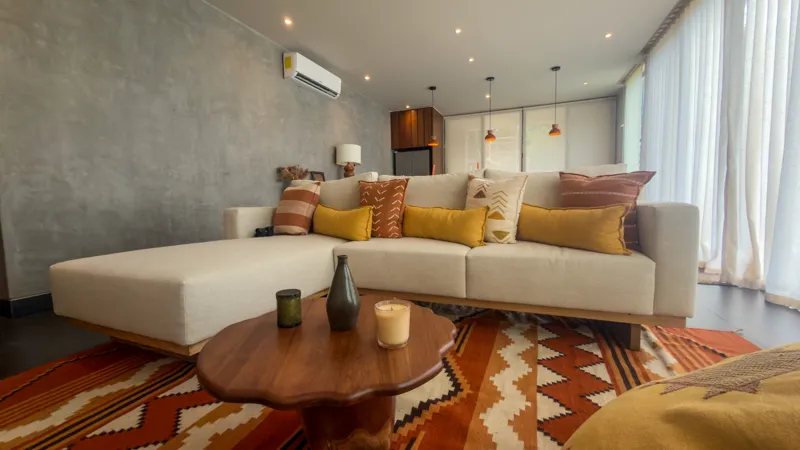 A wide shot of the open-concept dining area and kitchen in the Mestiza 15 showroom, featuring a long dark granite kitchen island with wooden bar stools, a wooden dining table set for six, modern pendant lights, and a contemporary kitchen with wood...