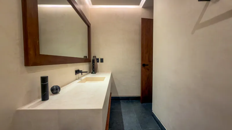 A wide shot of the open-concept dining area and kitchen in the Mestiza 15 showroom, featuring a long dark granite kitchen island with wooden bar stools, a wooden dining table set for six, modern pendant lights, and a contemporary kitchen with wood...