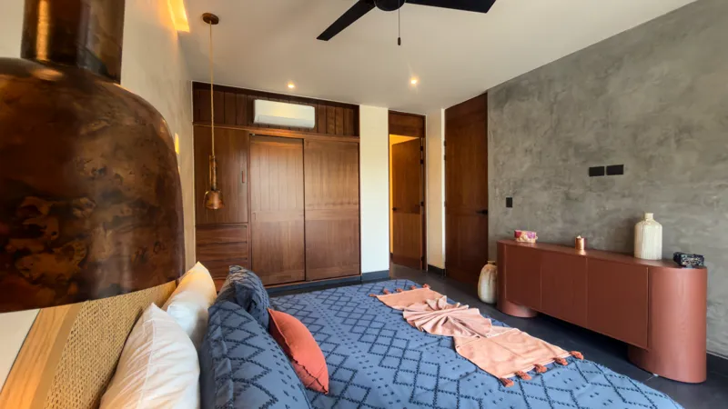 A wide shot of the open-concept dining area and kitchen in the Mestiza 15 showroom, featuring a long dark granite kitchen island with wooden bar stools, a wooden dining table set for six, modern pendant lights, and a contemporary kitchen with wood...