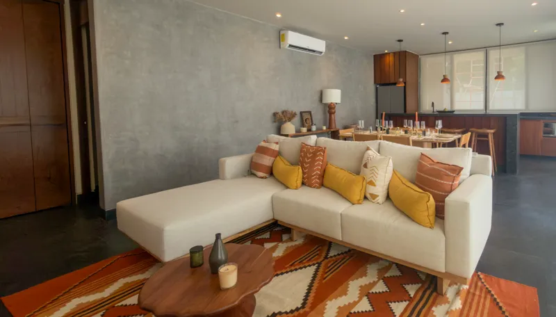 A wide shot of the open-concept dining area and kitchen in the Mestiza 15 showroom, featuring a long dark granite kitchen island with wooden bar stools, a wooden dining table set for six, modern pendant lights, and a contemporary kitchen with wood...