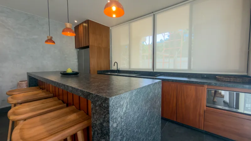 A wide shot of the open-concept dining area and kitchen in the Mestiza 15 showroom, featuring a long dark granite kitchen island with wooden bar stools, a wooden dining table set for six, modern pendant lights, and a contemporary kitchen with wood...