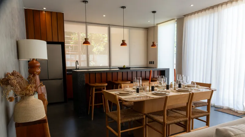 A wide shot of the open-concept dining area and kitchen in the Mestiza 15 showroom, featuring a long dark granite kitchen island with wooden bar stools, a wooden dining table set for six, modern pendant lights, and a contemporary kitchen with wood...