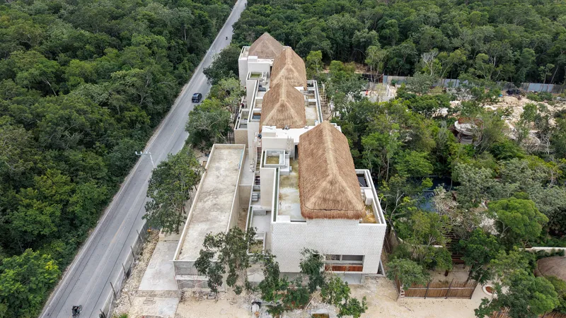An aerial view of the Mestiza 15 development in Tulum, showing a multi-story building with several thatched palapa roofs largely completed, surrounded by lush green jungle. To the left of the main building, new foundations and concrete structures ...