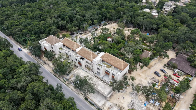 An aerial view of the Mestiza 15 development in Tulum, showing a multi-story building with several thatched palapa roofs largely completed, surrounded by lush green jungle. To the left of the main building, new foundations and concrete structures ...