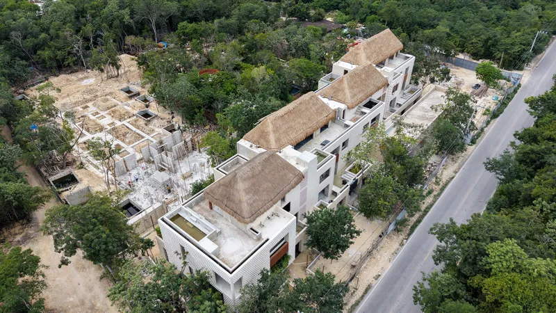 An aerial view of the Mestiza 15 development in Tulum, showing a multi-story building with several thatched palapa roofs largely completed, surrounded by lush green jungle. To the left of the main building, new foundations and concrete structures ...