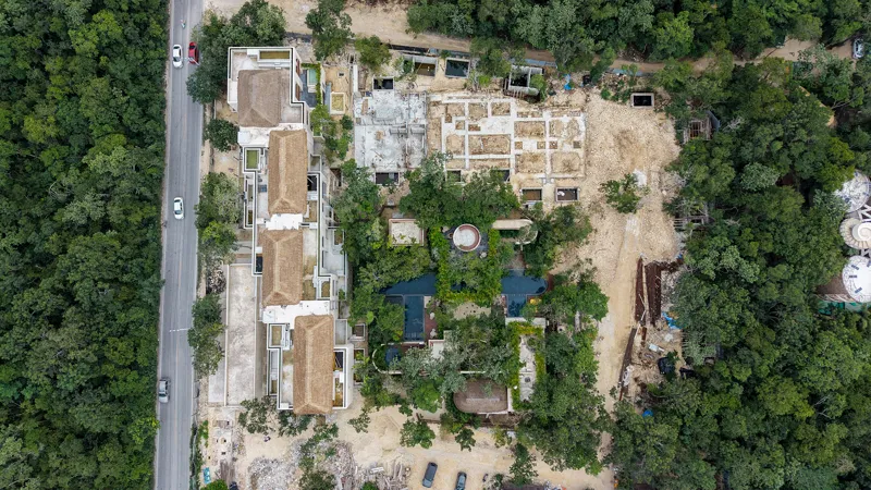 An aerial view of the Mestiza 15 development in Tulum, showing a multi-story building with several thatched palapa roofs largely completed, surrounded by lush green jungle. To the left of the main building, new foundations and concrete structures ...