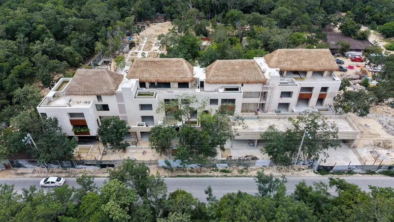 An aerial view of the Mestiza 15 development in Tulum, showing a multi-story building with several thatched palapa roofs largely completed, surrounded by lush green jungle. To the left of the main building, new foundations and concrete structures ...