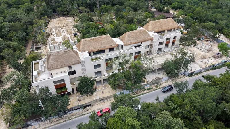 An aerial view of the Mestiza 15 development in Tulum, showing a multi-story building with several thatched palapa roofs largely completed, surrounded by lush green jungle. To the left of the main building, new foundations and concrete structures ...