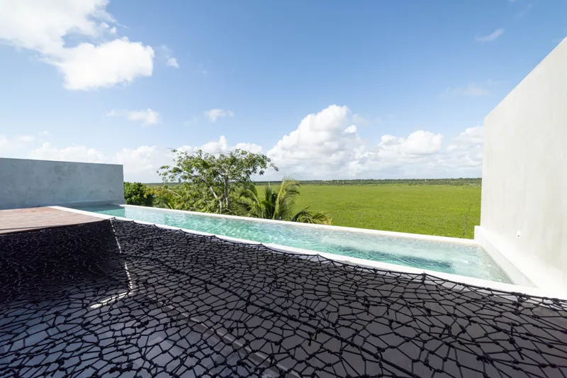 A breathtaking rooftop infinity pool at NEEA, overlooking a vibrant green jungle landscape under a bright blue sky, with a modern relaxation net area in the foreground.
