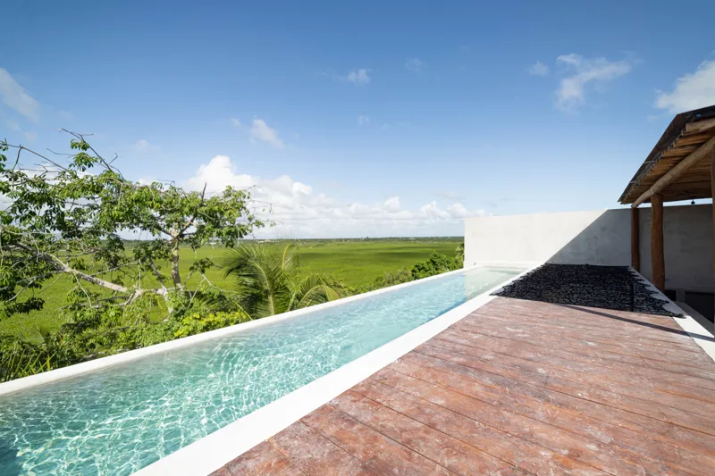 A breathtaking rooftop infinity pool at NEEA, overlooking a vibrant green jungle landscape under a bright blue sky, with a modern relaxation net area in the foreground.