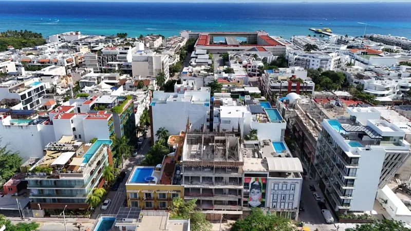 Aerial view of Distrito Playa under construction in Playa del Carmen, showing the multi-story building's concrete structure and framed floors, surrounded by other urban buildings and with the turquoise Caribbean Sea visible in the background.