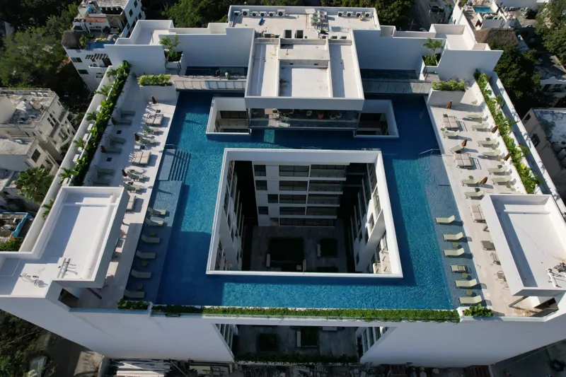 Aerial view of the Ceiba at 25 development in Playa del Carmen, Mexico, showing the multi-story white building with its completed rooftop pools and lounge areas, surrounded by other urban buildings, lush green trees, a sports stadium, and the turq...