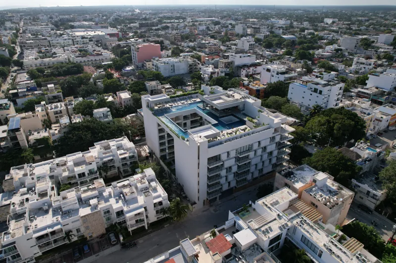 Aerial view of the Ceiba at 25 development in Playa del Carmen, Mexico, showing the multi-story white building with its completed rooftop pools and lounge areas, surrounded by other urban buildings, lush green trees, a sports stadium, and the turq...