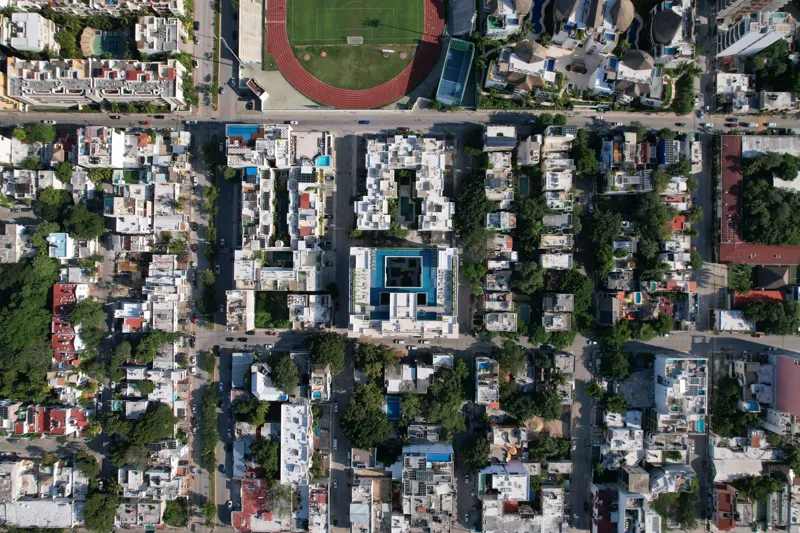 Aerial view of the Ceiba at 25 development in Playa del Carmen, Mexico, showing the multi-story white building with its completed rooftop pools and lounge areas, surrounded by other urban buildings, lush green trees, a sports stadium, and the turq...