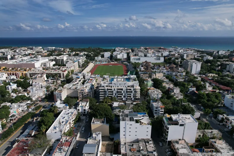Aerial view of the Ceiba at 25 development in Playa del Carmen, Mexico, showing the multi-story white building with its completed rooftop pools and lounge areas, surrounded by other urban buildings, lush green trees, a sports stadium, and the turq...
