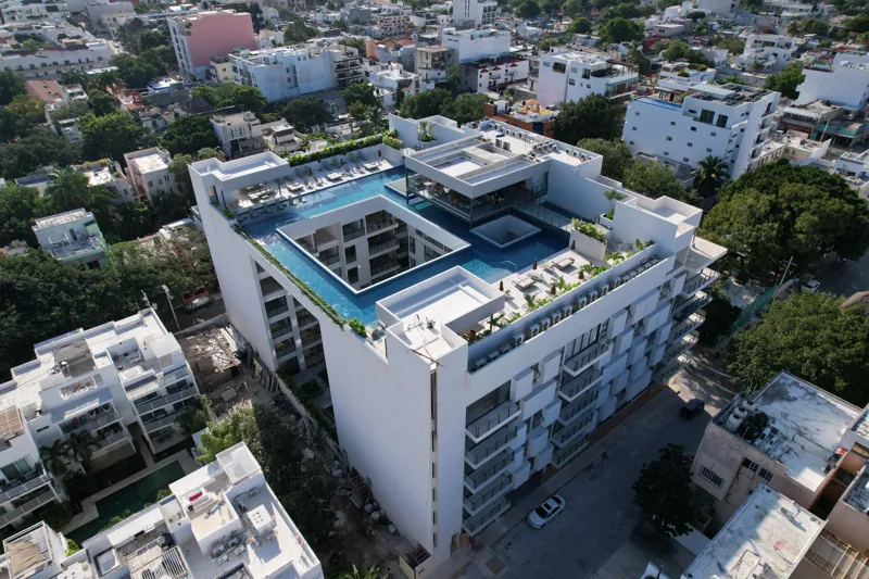Aerial view of the Ceiba at 25 development in Playa del Carmen, Mexico, showing the multi-story white building with its completed rooftop pools and lounge areas, surrounded by other urban buildings, lush green trees, a sports stadium, and the turq...