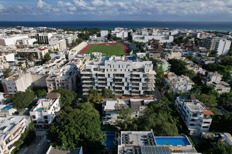 Aerial view of the Ceiba at 25 development in Playa del Carmen, Mexico, showing the multi-story white building with its completed rooftop pools and lounge areas, surrounded by other urban buildings, lush green trees, a sports stadium, and the turq...