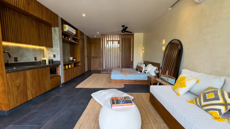 A wide shot of a luxurious studio apartment in Mestiza 15, featuring a modern kitchenette with dark countertops and wooden cabinetry, a sleeping area with a bed separated by a wooden slat partition, and a living space with a light-colored sofa, al...