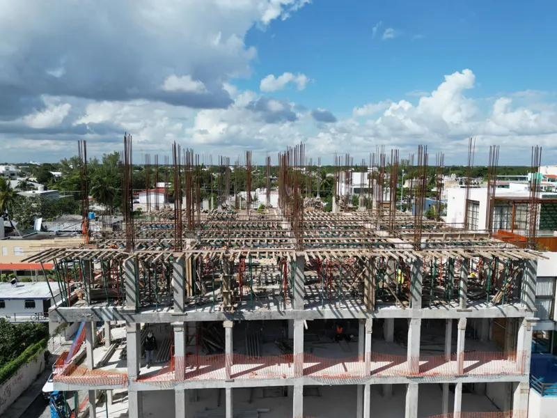 Aerial view of the Maia development construction site, showing the concrete structure of multiple floors with extensive wooden formwork and rebar for upcoming levels, surrounded by lush green trees and a construction vehicle on the ground.