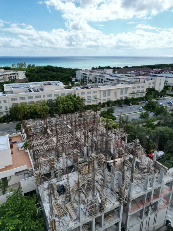 Aerial view of the Maia development construction site, showing the concrete structure of multiple floors with extensive wooden formwork and rebar for upcoming levels, surrounded by lush green trees and a construction vehicle on the ground.