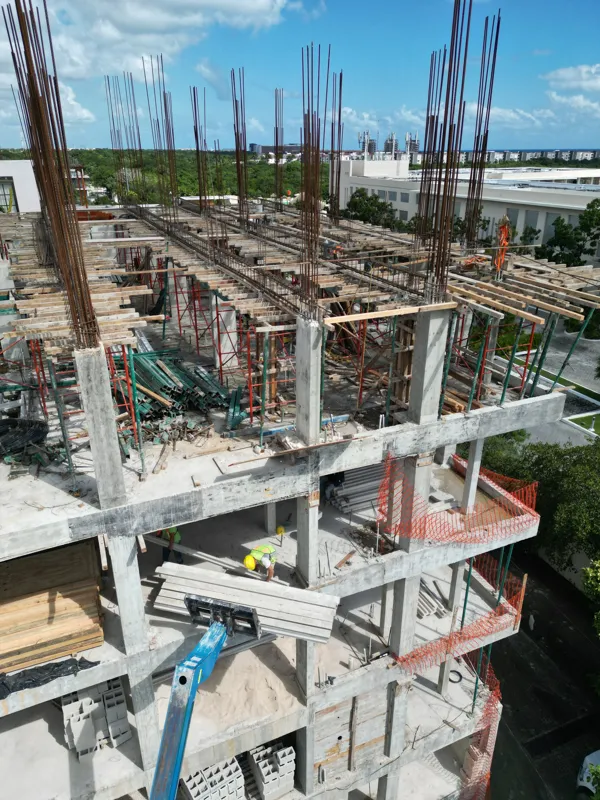 Aerial view of the Maia development construction site, showing the concrete structure of multiple floors with extensive wooden formwork and rebar for upcoming levels, surrounded by lush green trees and a construction vehicle on the ground.
