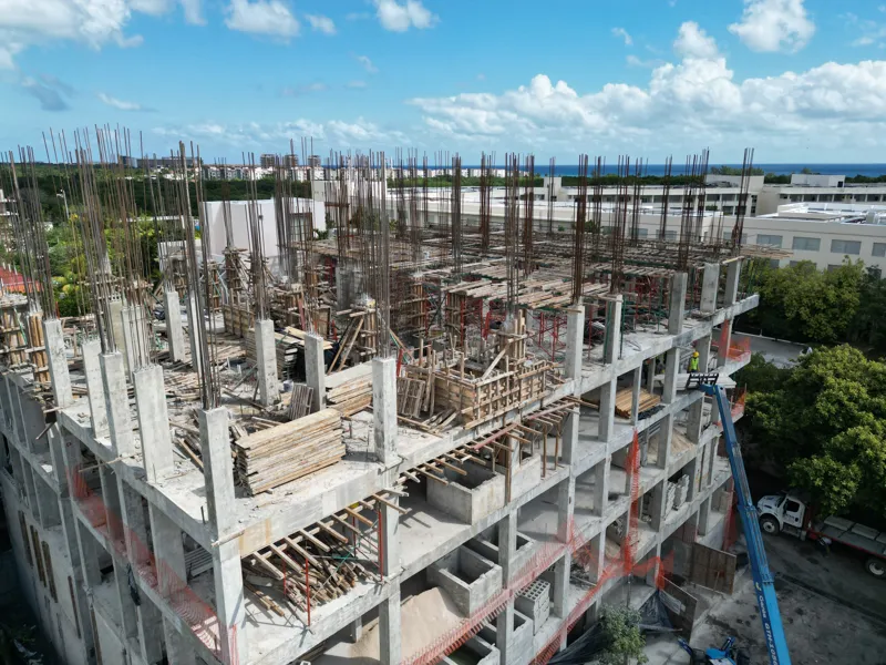 Aerial view of the Maia development construction site, showing the concrete structure of multiple floors with extensive wooden formwork and rebar for upcoming levels, surrounded by lush green trees and a construction vehicle on the ground.