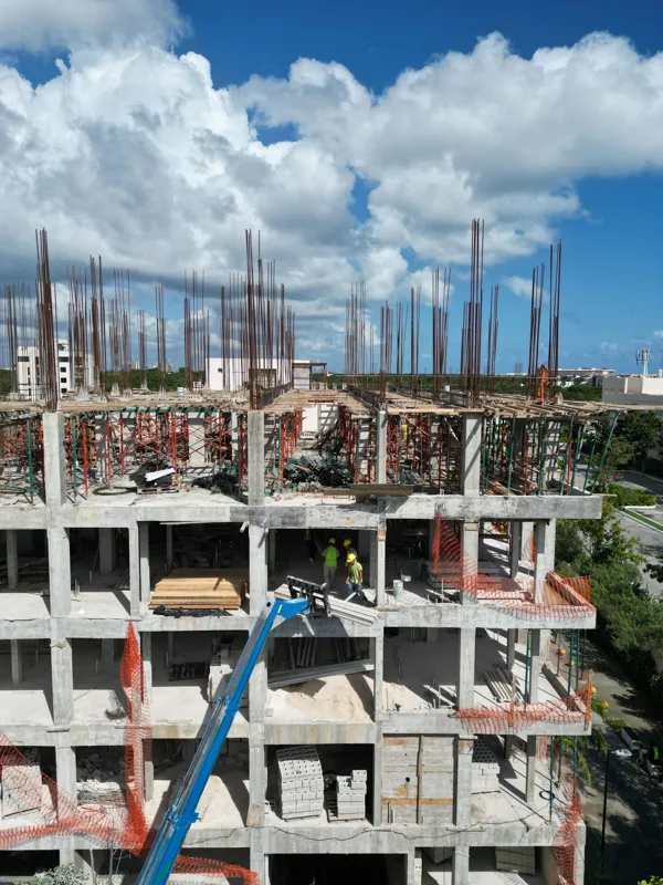 Aerial view of the Maia development construction site, showing the concrete structure of multiple floors with extensive wooden formwork and rebar for upcoming levels, surrounded by lush green trees and a construction vehicle on the ground.