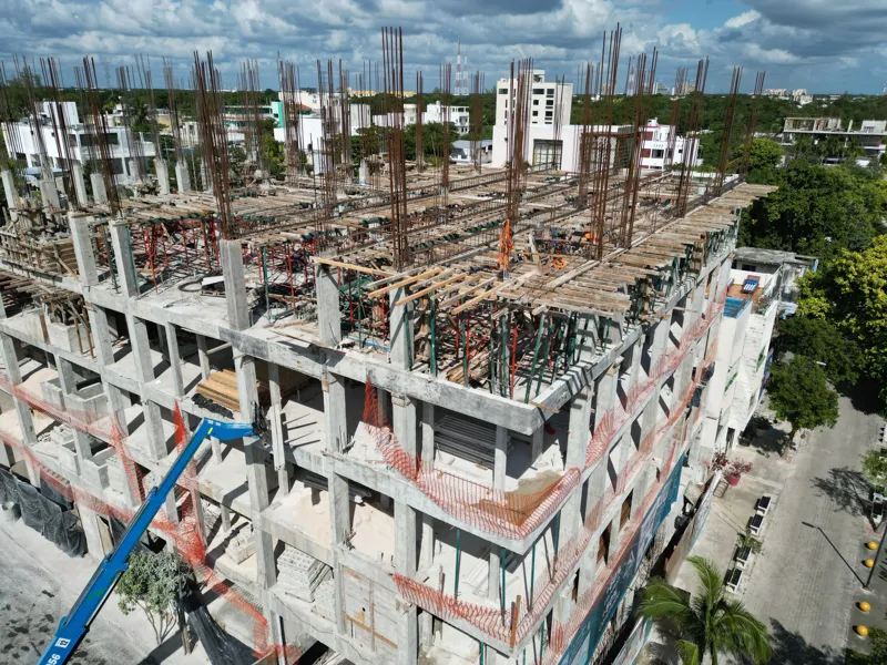Aerial view of the Maia development construction site, showing the concrete structure of multiple floors with extensive wooden formwork and rebar for upcoming levels, surrounded by lush green trees and a construction vehicle on the ground.