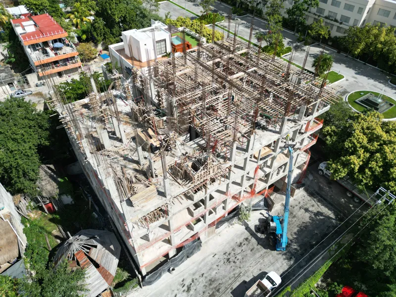 Aerial view of the Maia development construction site, showing the concrete structure of multiple floors with extensive wooden formwork and rebar for upcoming levels, surrounded by lush green trees and a construction vehicle on the ground.