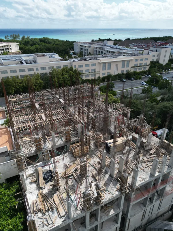 Aerial view of the Maia development construction site, showing the concrete structure of multiple floors with extensive wooden formwork and rebar for upcoming levels, surrounded by lush green trees and a construction vehicle on the ground.