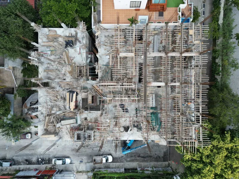 Aerial view of the Maia development construction site, showing the concrete structure of multiple floors with extensive wooden formwork and rebar for upcoming levels, surrounded by lush green trees and a construction vehicle on the ground.