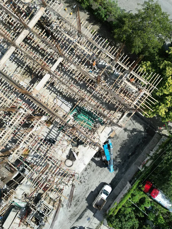 Aerial view of the Maia development construction site, showing the concrete structure of multiple floors with extensive wooden formwork and rebar for upcoming levels, surrounded by lush green trees and a construction vehicle on the ground.