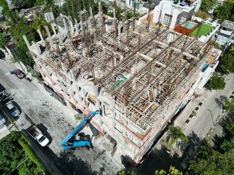 Aerial view of the Maia development construction site, showing the concrete structure of multiple floors with extensive wooden formwork and rebar for upcoming levels, surrounded by lush green trees and a construction vehicle on the ground.