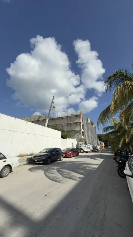 Exterior view of the Costa Celeste apartment complex under construction in Playa del Carmen, January 2026. The multi-story concrete structure is partially covered with black safety netting and scaffolding, indicating ongoing work. Cars are parked ...