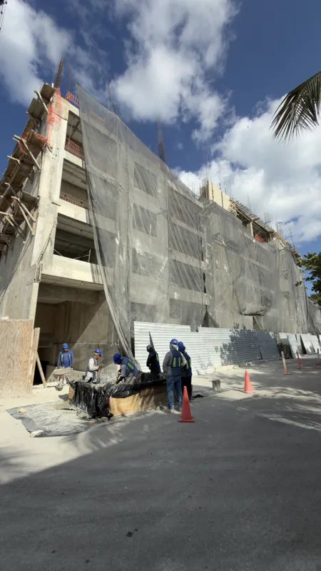 Exterior view of the Costa Celeste apartment complex under construction in Playa del Carmen, January 2026. The multi-story concrete structure is partially covered with black safety netting and scaffolding, indicating ongoing work. Cars are parked ...