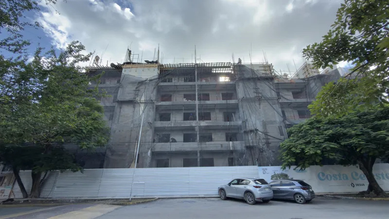 Exterior view of the Costa Celeste apartment complex under construction in Playa del Carmen, January 2026. The multi-story concrete structure is partially covered with black safety netting and scaffolding, indicating ongoing work. Cars are parked ...