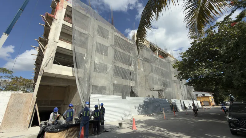 Exterior view of the Costa Celeste apartment complex under construction in Playa del Carmen, January 2026. The multi-story concrete structure is partially covered with black safety netting and scaffolding, indicating ongoing work. Cars are parked ...