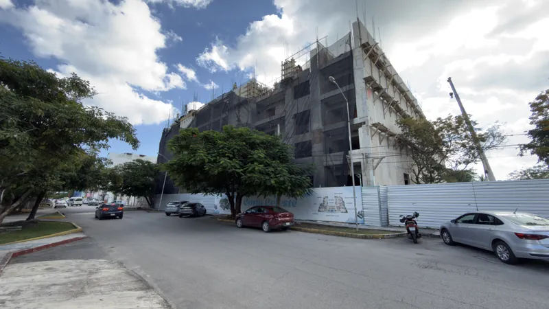 Exterior view of the Costa Celeste apartment complex under construction in Playa del Carmen, January 2026. The multi-story concrete structure is partially covered with black safety netting and scaffolding, indicating ongoing work. Cars are parked ...