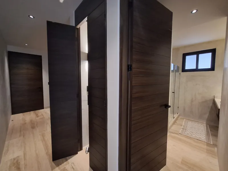 A modern kitchen and dining area in an Attha Holistika 1-bedroom unit, featuring light wood cabinetry, a stainless steel refrigerator, a black countertop, a round wooden dining table with four chairs, and a ceiling fan, with a large window offerin...