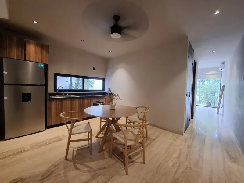 A modern kitchen and dining area in an Attha Holistika 1-bedroom unit, featuring light wood cabinetry, a stainless steel refrigerator, a black countertop, a round wooden dining table with four chairs, and a ceiling fan, with a large window offerin...