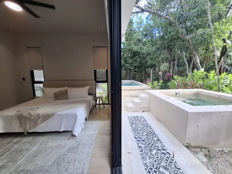 A modern kitchen and dining area in an Attha Holistika 1-bedroom unit, featuring light wood cabinetry, a stainless steel refrigerator, a black countertop, a round wooden dining table with four chairs, and a ceiling fan, with a large window offerin...