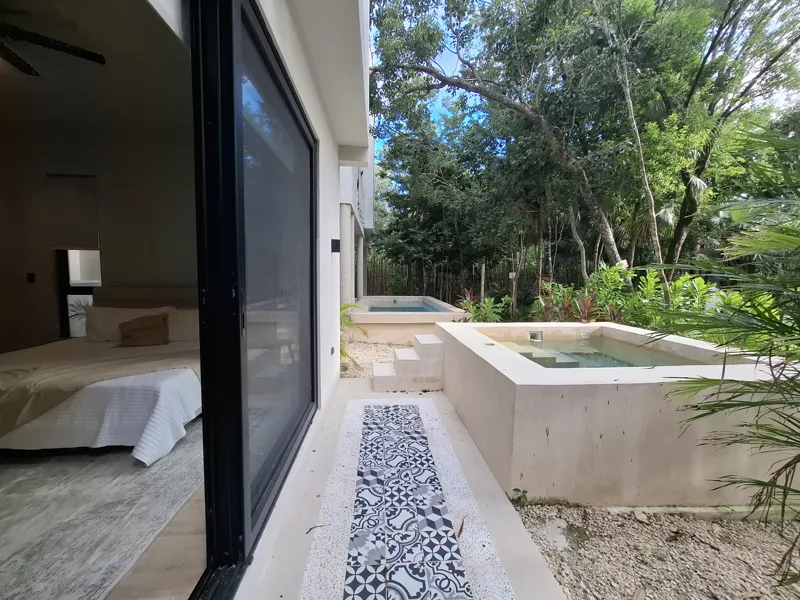 A modern kitchen and dining area in an Attha Holistika 1-bedroom unit, featuring light wood cabinetry, a stainless steel refrigerator, a black countertop, a round wooden dining table with four chairs, and a ceiling fan, with a large window offerin...