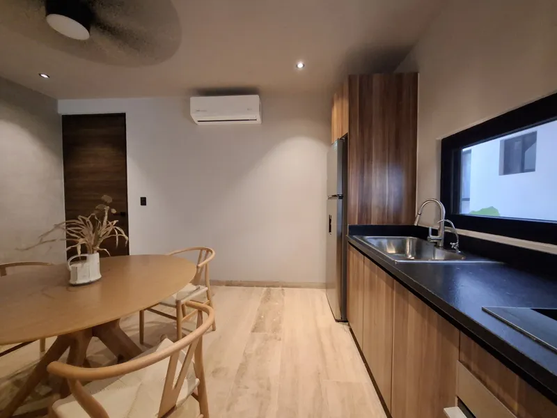 A modern kitchen and dining area in an Attha Holistika 1-bedroom unit, featuring light wood cabinetry, a stainless steel refrigerator, a black countertop, a round wooden dining table with four chairs, and a ceiling fan, with a large window offerin...