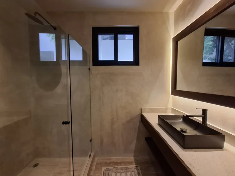 A modern kitchen and dining area in an Attha Holistika 1-bedroom unit, featuring light wood cabinetry, a stainless steel refrigerator, a black countertop, a round wooden dining table with four chairs, and a ceiling fan, with a large window offerin...