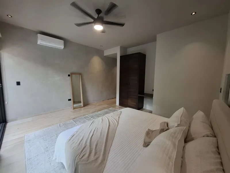 A modern kitchen and dining area in an Attha Holistika 1-bedroom unit, featuring light wood cabinetry, a stainless steel refrigerator, a black countertop, a round wooden dining table with four chairs, and a ceiling fan, with a large window offerin...