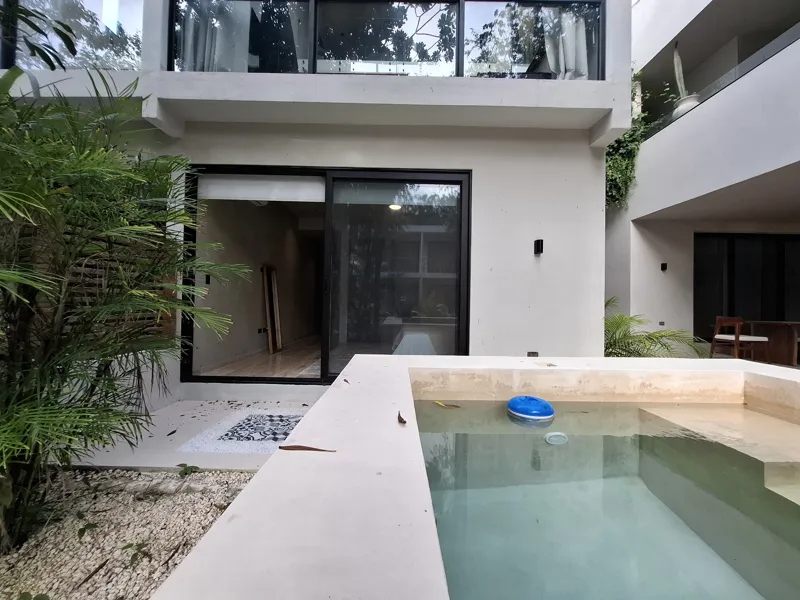 A modern kitchen and dining area in an Attha Holistika 1-bedroom unit, featuring light wood cabinetry, a stainless steel refrigerator, a black countertop, a round wooden dining table with four chairs, and a ceiling fan, with a large window offerin...