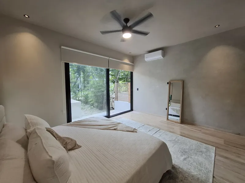 A modern kitchen and dining area in an Attha Holistika 1-bedroom unit, featuring light wood cabinetry, a stainless steel refrigerator, a black countertop, a round wooden dining table with four chairs, and a ceiling fan, with a large window offerin...