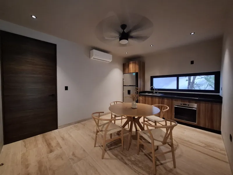 A modern kitchen and dining area in an Attha Holistika 1-bedroom unit, featuring light wood cabinetry, a stainless steel refrigerator, a black countertop, a round wooden dining table with four chairs, and a ceiling fan, with a large window offerin...